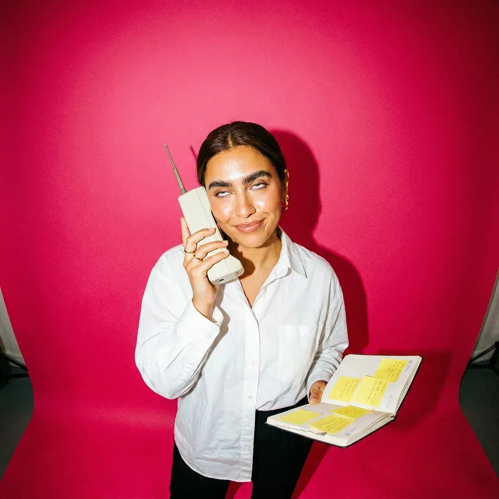 Woman on a retro brick phone, rolling her eyes, holding a diary full of sticky notes, on a hot pink backdrop