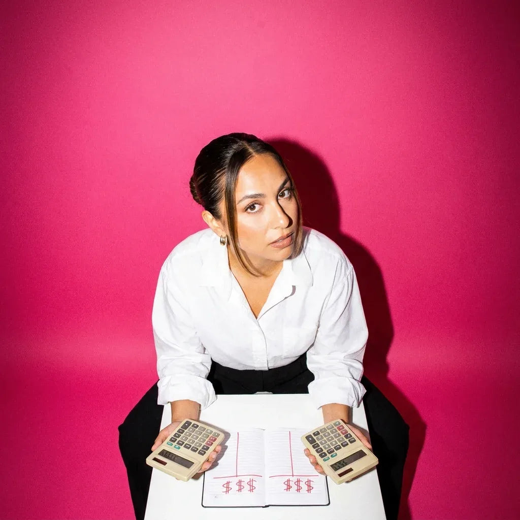 Woman at a white desk holding two retro calculators over a notebook with two columns of dollar signs